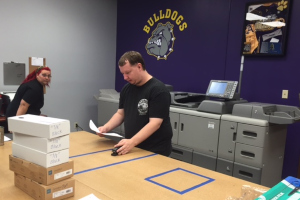 Primary: Man stapling papers together, Secondary: large printer and woman watching, Background: Ark City Bulldogs logo painted on the wall